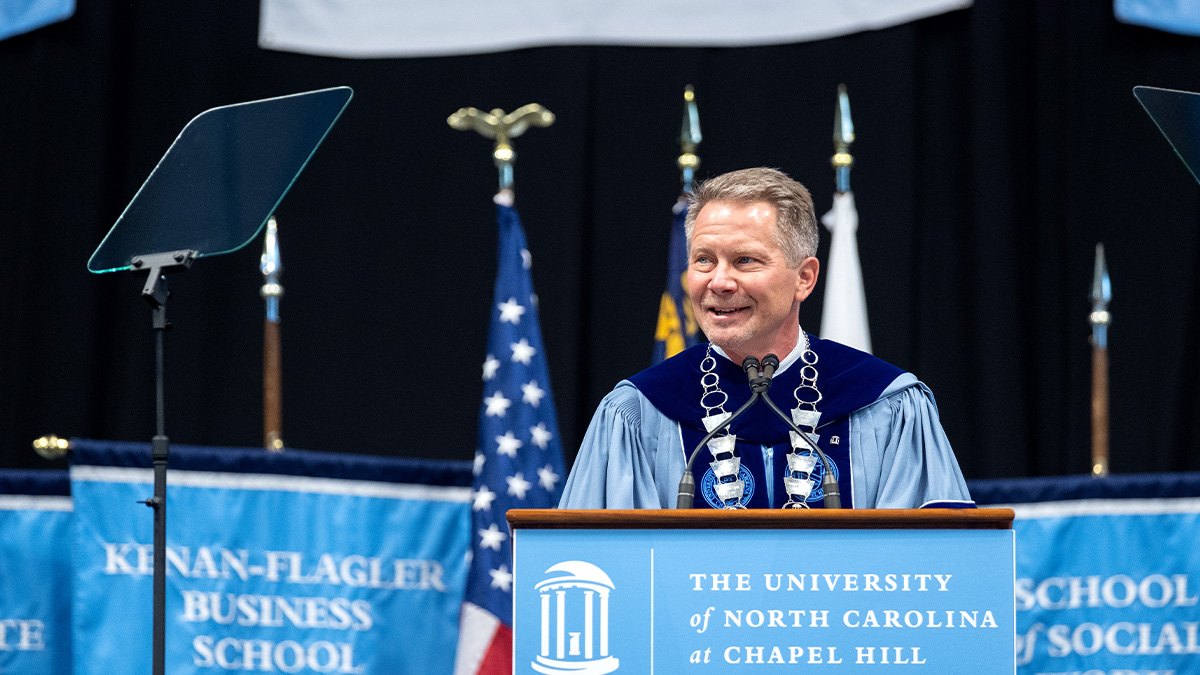 Kevin M. Guskiewicz wearing regalia and smiling while speaking from a podium at Winter Commencement.