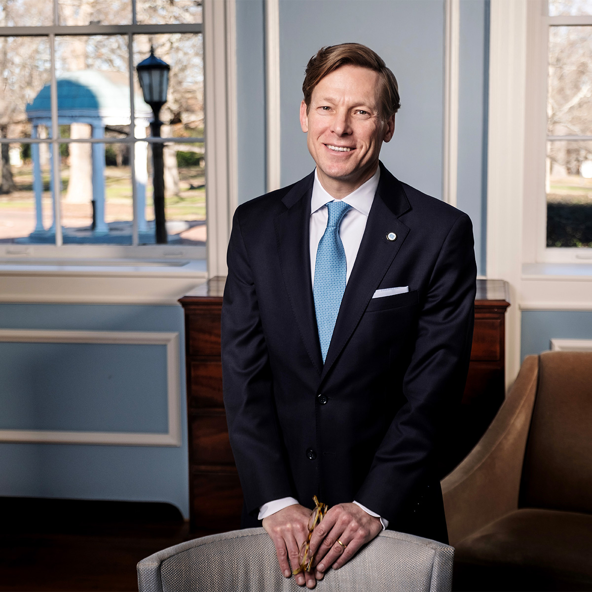 A man, UNC-Chapel Hill interim chancellor Lee H. Roberts, posing for a photo in an office in South Building with the Old Well seen through a window in the background.